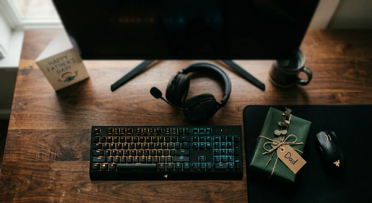 Warm flat lay of gaming peripherals, a gift box with "Dad" tag, and a Father's Day card on a dark desk.